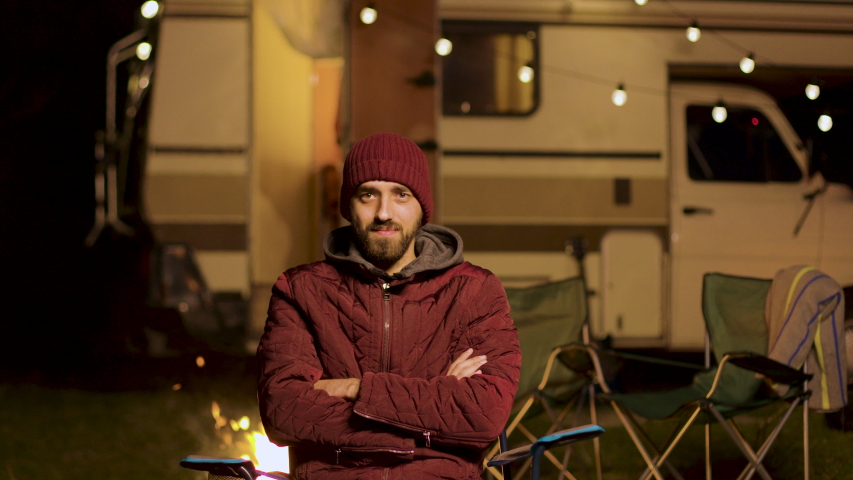 Portrait of bearded young man sitting on a chair looking at the camera. Retro camper van. Light bulbs.