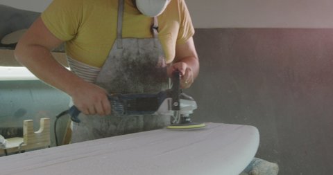 Front view of a millennial Caucasian male surfboard maker in his workshop wearing a face mask and polishing a new surfboard with a power tool during the manufacturing process