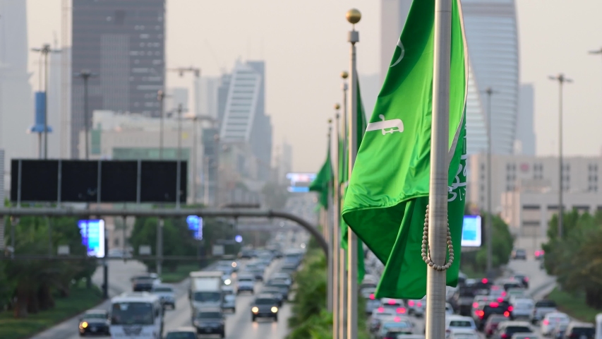 Saudi Arabia flag waving and Riyadh's main street on background. 