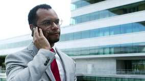 Focused businessman talking by smartphone. Side view of young African American businessman in formal wear talking by cell phone and walking outside modern office building. Communication concept