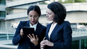 Focused businesswomen with digital devices. Serious multiethnic businesswomen in formal wear standing together on street and using digital tablet. Technology concept
