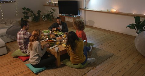 High angle view of a group of young adult multi-ethnic male and female friends sitting at a low table at home set for Thanksgiving dinner, eating and talking