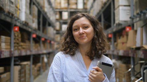 An attractive girl with curly hair in a shirt stands with a tablet in her hands in a warehouse between the shelves with goods, smiles and looks at the camera in slow motion, close-up portrait