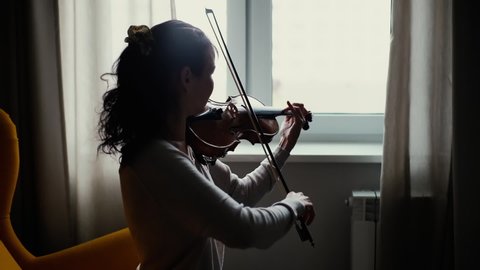 Beautiful young woman musician playing the violin by the window, sitting on soft chair in room with a modern interior. Girl is practicing playing musical instrument at home.