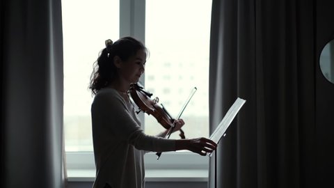Beautiful young woman violinist plays the violin at home by the window in slow motion. Girl musician looks at the score.