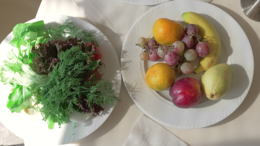 A top view of three white plates full of healthy food. Fruits, greens and vegetables. A woman taking some tomatoes from one of the plates. 