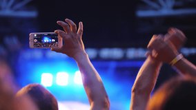 A slow motion of two men in the crowd enjoying an open air concert show. One of them is holding a smartphone and taking video of a faraway stage. Another guy is waving his arms above the head