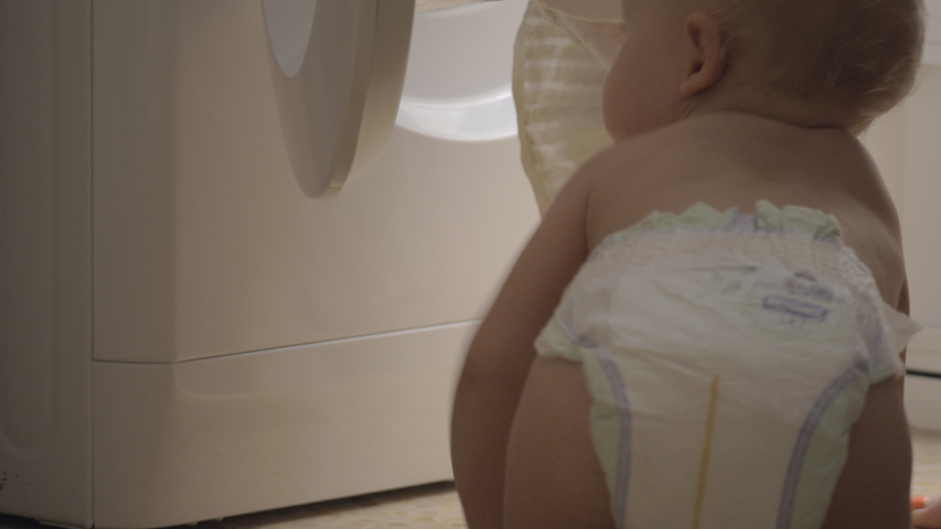 One year old baby girl is looking at mother putting bed linen into the washing machine