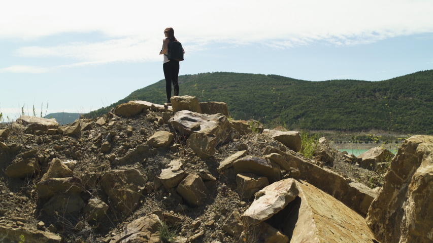 Attractive explorer redhead girl standing hiking at beautiful blue turquoise lake of Yesa, black backpack, with rocks and green forest background during sunny summer in Spain. Wide Crane shot 4K UHD.
