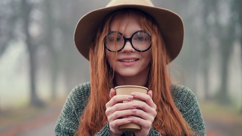 Close up portrait of young woman in hat and glasses drinking hot coffee in the autumn park. Slowmotion.