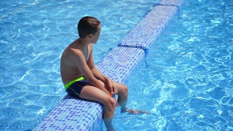 Boy having a rest in the pool. Active boy sitting on the swimming pool after swimming on blue clear water background outdoors. Healthy concept.