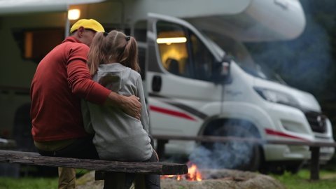 RV Campsite Family Time. Father Hugging His Daughter on Wooden Bench in Front of Campfire and Motorhome.