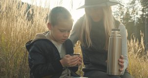 Happy mother and son hiking traveler camper drinking hot tea and relaxing in fall forest on sunny autumn day. Beautiful mountain landscape view. 4K slow motion video.