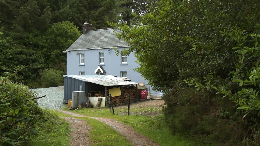 Wide shot of a two story blue farmhouse tucked away behind trees in a forest, with a dirt driveway leading to a gravel parking lot near a firewood shed
