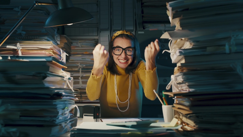 Happy cheerful woman celebrating in the office with fists raised, she is surrounded by piles of paperwork | Shutterstock HD Video #1040314406