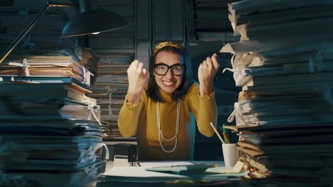 Happy cheerful woman celebrating in the office with fists raised, she is surrounded by piles of paperwork