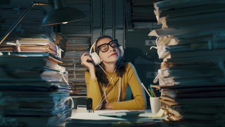 Happy cheerful secretary wearing headphones and listening to music in the office instead of working | Shutterstock HD Video #1040314427
