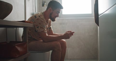 In the bathroom young man on toilet relaxing playing mobile game on smartphone winning cheering up. Fun time.