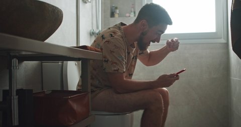 Funny-looking young bearded man sitting on toilet browsing smartphone rejoicing with financial success or profits clenching fist staying in the restroom.