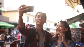 Young happy smiling Asian tourist couple taking smartphone selfies while walking along Khao San Road market Bangkok Thailand at sunset in slow motion with lens flare