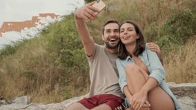 Attractive cheerful young lovely couple hugging and taking selfie photo on smartphone while sitting outdoors on green hills