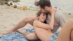 Handsome calm young bearded man hugging with his cute young girlfriend and kissing her in forehead while lying on plaid near the ocean
