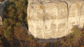 Aerial fly around a young blonde female hiker with backpack with raised hands up, feeling freedom while standing at the edge of stunning rock formation surrounded by autumn forest
