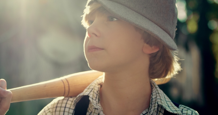 Portrait of the little Caucasian cute teen hooligan boy in a hat posing to the camera and holding a bat over his shoulder. Close up. Outdoor.