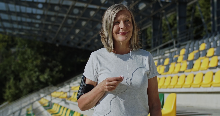 Happy Caucasian senior sporty woman putting headphones in ears as listening to the music and doing sport at the stadium. Outside.