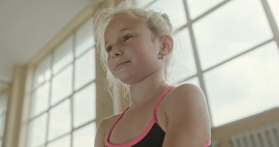 Low angle portrait of a smiling young blond ballerina in a dance studio looking ahead against bright windows