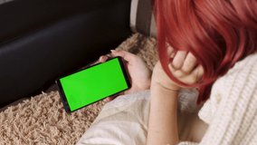 teenager girl on a sofa, watches video on her phone, clicks on the screen. Mock-up with green screen for tracking close-up. Social media and device technology. Woman smartphone greenscreen frame.