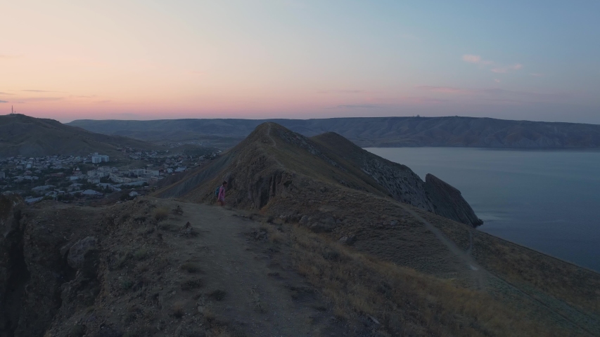 Aerial view young tanned girl with a backpack coming down from the mountain. Cityscape and mountains during sunset in a background