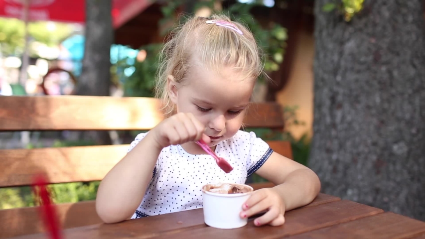 Aborable smiling blonde 3 years baby girl eating broun ice cream in the park at summer day and laughing