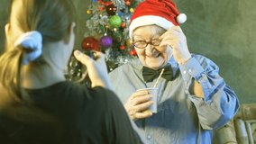young girl with large professional video camera photographs elderly woman. Atractive pensioner in bow-tie and Santa Claus hat is holding eco-friendly cardboard glass with straw. Studio shooting elder