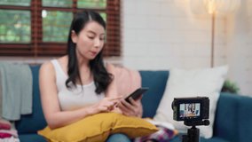 Asian fashion influencer designer women using camera streaming and live to sell clothes business, e-commerce broadcast online using mobile phone chat answer customer on sofa in living room at home.