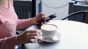 Woman using smartphone and drinking coffee at cafe table outdoor, slow motion video. Girl scrolls her news feed on smartphone during lunch break on restaurant terrace, close up