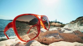 Close-up view of red fashionable sunglasses on sea stone beach. Rock coast on summer happy day. Rays lights on background. Vacation abroad. Date meeting website online. Business weekend.