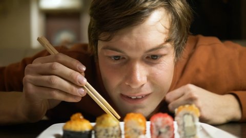 man smacking lips and looking at plate with desire eats sushi rolls of different types with wooden chopsticks closeup