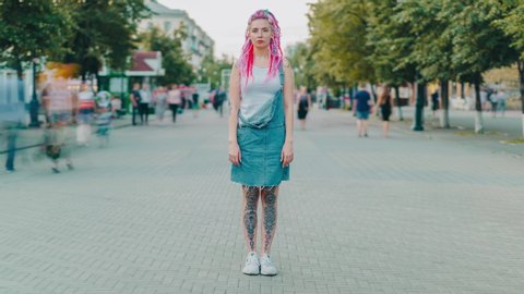 Time lapse portrait of cool young woman with colorful hair and tattoo standing in city street while people are moving around. Youth and modern style concept.