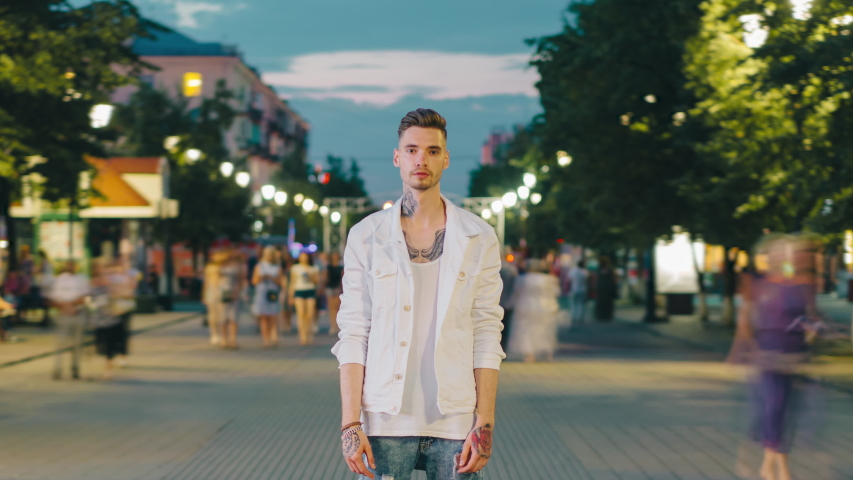 Time lapse portrait of tattoed student standing in urban pedestrian street at night looking at camera with serious expression. People and lifestyle concept.