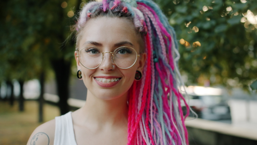 Close-up portrait of joyful young woman with bright hair, piercing and dental braces standing alone in park smiling looking at camera. People and style concept.