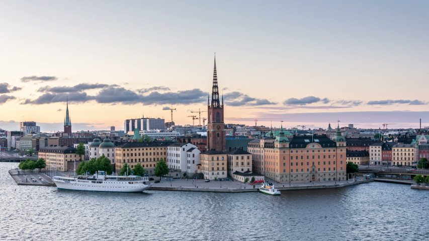 Stockholm city skyline at dusk. Riddarholmen islet with Riddarholmen church tower, part of Gamla Stan. Clouds move fast across the purple sunset sky. City lights switch on. Time lapse video.