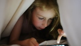 Little girl is playing game on the phone under the covers.