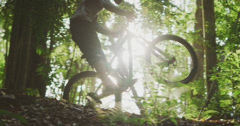 Cinematic slow motion shot of an extreme mountain biker riding downhill doing a stylish manual silhouetted in the afternoon sun surrounded by green forest, summer fun outdoors mountain biking
