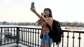 Young cute smiling Asian woman tourist with backpack taking selfie with smartphone and waving on river wharf at sunset in Bangkok Thailand