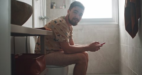 Bearded young caucasian man using smartphone playing mobile game while sitting on toilet in the bathroom. Fun home scene.