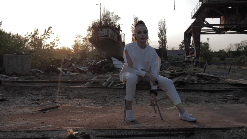 young woman in sweater with open sleeves sits on chair against damaged metal port parts and sundown slow motion