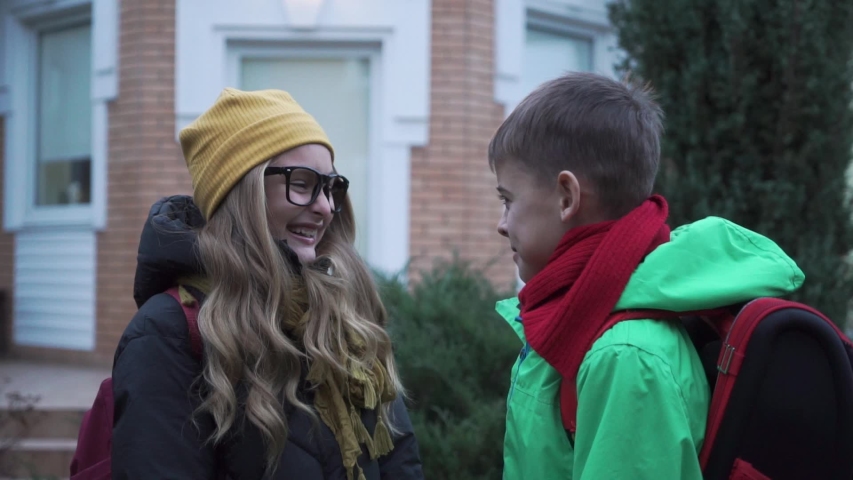 Close-up of Caucasian schoolmates laughing and hugging next to the big house. Positive boy and girl with backpacks leaving to school. Siblings enjoying morning before classes.