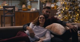 Caucasian couple sitting together on a sofa and watching TV at home during Christmas holidays, decorated apartment interior. 4K UHD RAW graded footage
