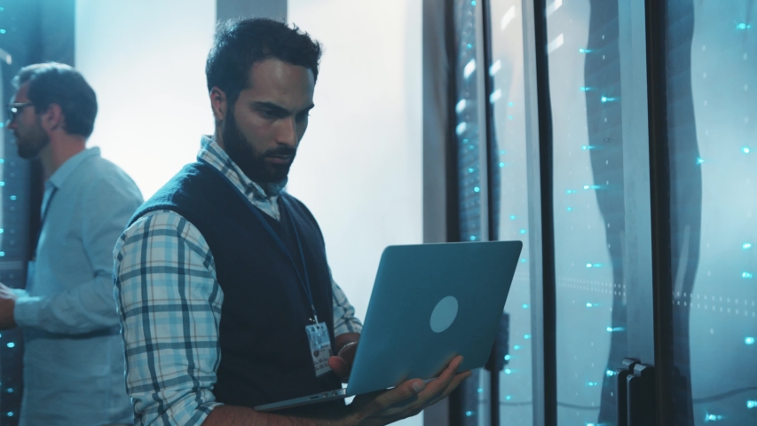 Data center male specialists cooperating in server room. Bearded IT admin activating computer hardware in rack cabinets behing glass panels. Webhosting. Technology concept.